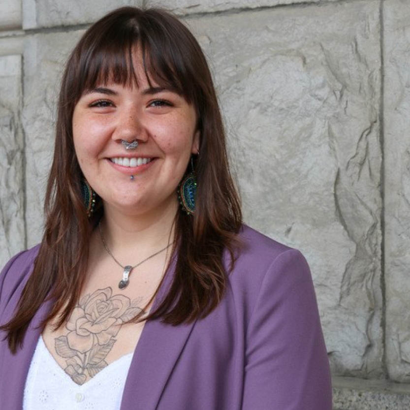 A smiling young woman with a nose ring and tatoo in a suit jacket.